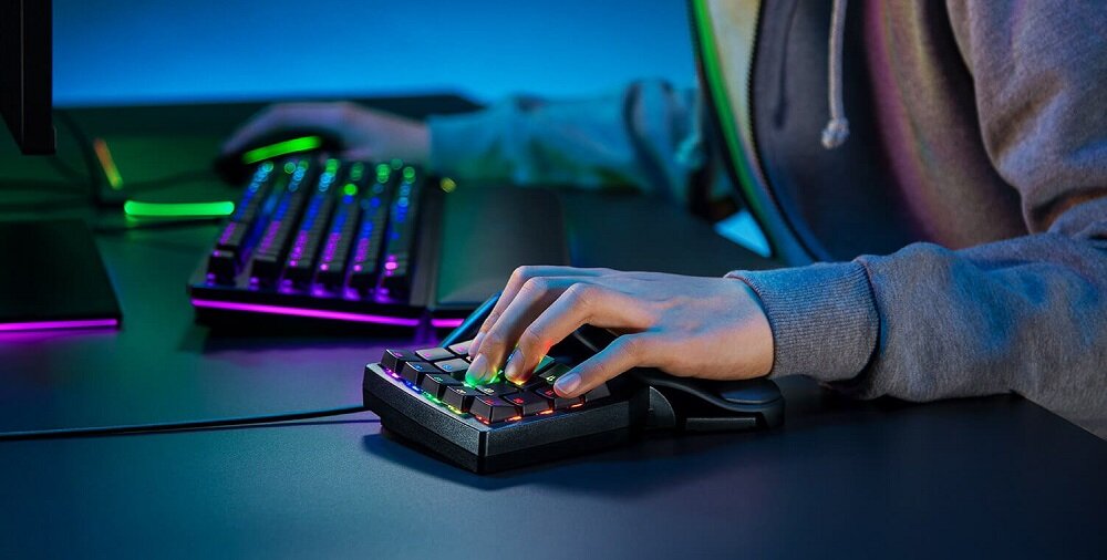 Osoba używa klawiatury z podświetleniem RGB, obok znajduje się pełnowymiarowa klawiatura również z podświetleniem. Obie klawiatury znajdują się na ciemnym biurku.