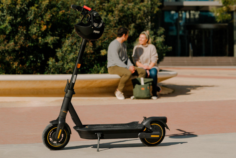 Czarna hulajnoga elektryczna stoi na chodniku, w tle widać dwie osoby siedzące na ławce. Na kierownicy hulajnogi znajduje się kask z widocznym logo \'Segway\'.