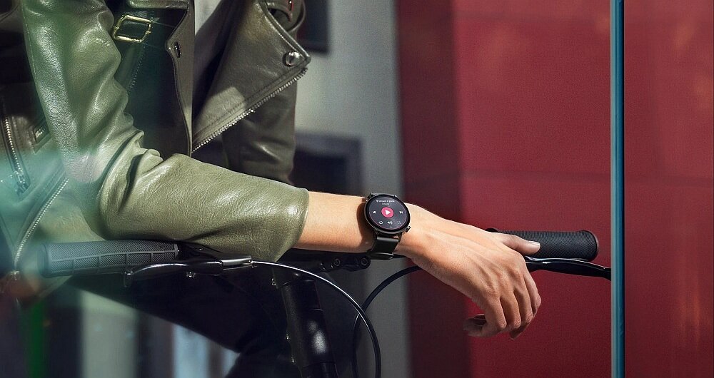 Osoba w zielonej kurtce trzyma kierownicę roweru, na nadgarstku ma czarny smartwatch z okrągłym wyświetlaczem. Na ekranie zegarka widoczne są ikony i czerwony przycisk.