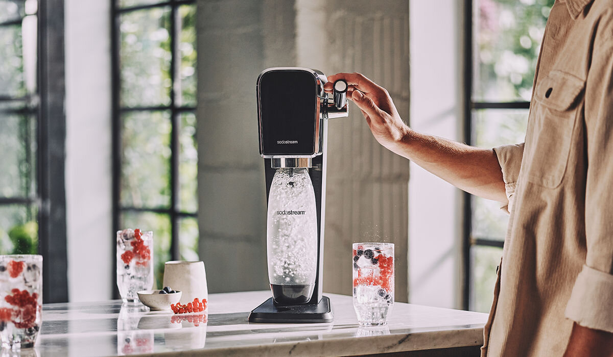 Na blacie kuchennym stoi czarny saturator z logo \'sodastream\', w którym znajduje się butelka z wodą gazowaną. Obok widoczne są szklanki z napojem i owocami oraz miseczki z dodatkami.