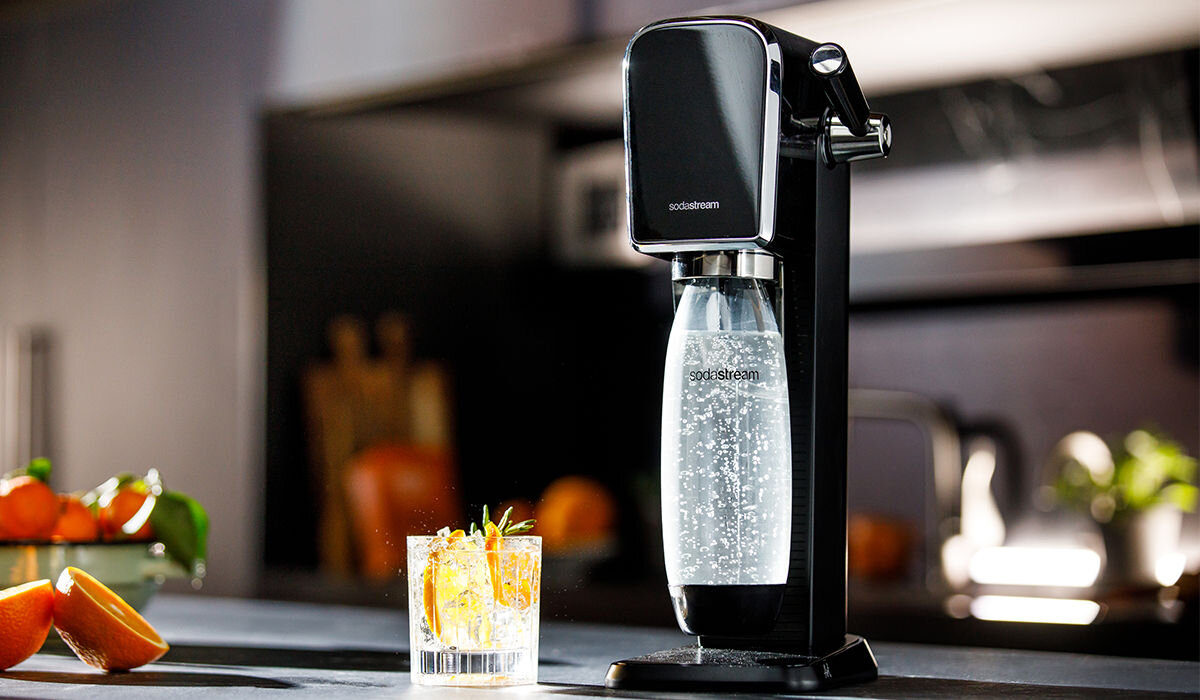 Na blacie kuchennym stoi czarny saturator z logo \'sodastream\', obok którego znajduje się szklanka z napojem i plasterkami pomarańczy. W tle widoczne są rozmyte elementy kuchni oraz owoce.