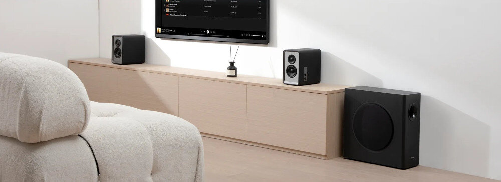 Na obrazie widać nowoczesne wnętrze z jasną sofą, szafką RTV i sprzętem audio. Po prawej stronie znajduje się czarny subwoofer, a na szafce stoją dwa głośniki i telewizor z wyświetlonym interfejsem aplikacji muzycznej.