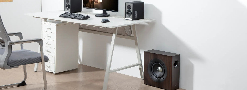 Na zdjęciu znajduje się biurko z komputerem, klawiaturą i myszką, a obok niego stoi subwoofer w kolorze czarnym. W tle widoczne są głośniki na biurku oraz krzesło biurowe.