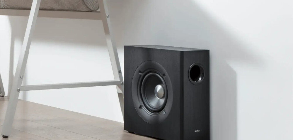 Czarny subwoofer z okrągłym głośnikiem i otworem bass-reflex stoi na drewnianej podłodze obok białej ściany. Obok znajduje się fragment metalowej drabinki.