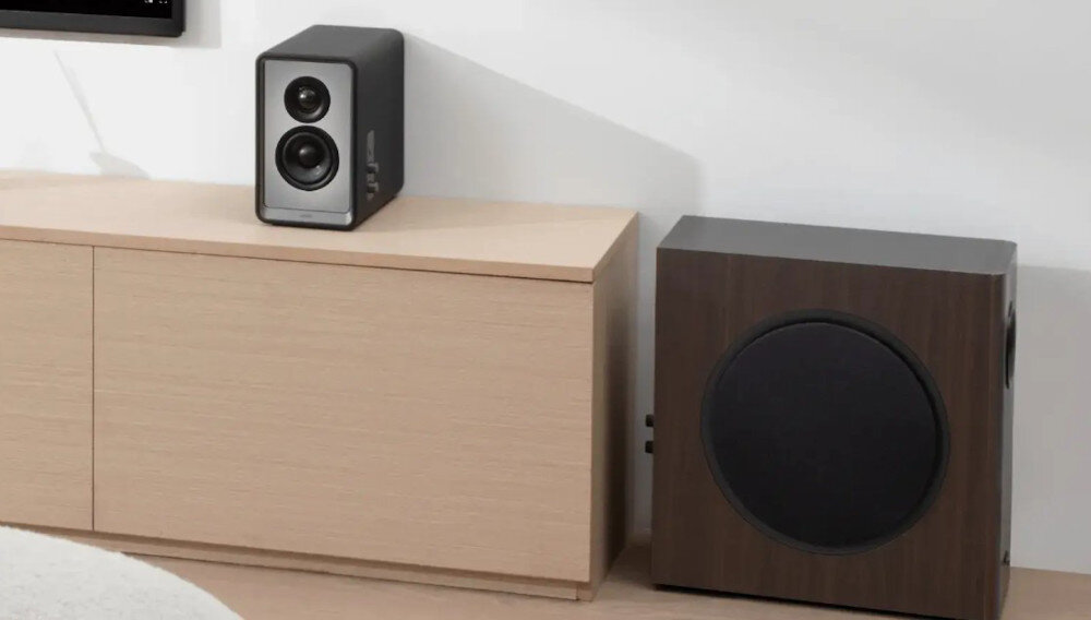 Na obrazie widoczny jest czarny subwoofer o prostokątnej obudowie z MDF oraz głośnik z dwoma przetwornikami umieszczony na jasnej szafce. Oba urządzenia mają nowoczesny design i znajdują się w minimalistycznym wnętrzu.
