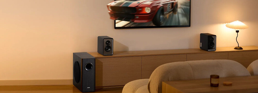 W salonie na szafce stoi telewizor z wyświetlonym obrazem samochodu oraz dwa głośniki. Obok szafki znajduje się subwoofer, a na niej lampa i dekoracyjny przedmiot.