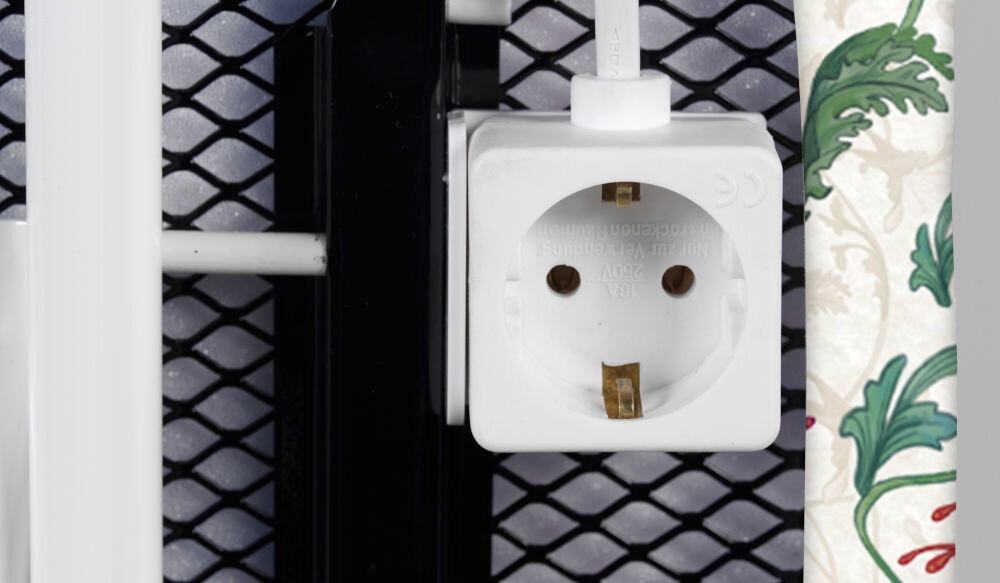 Zbliżenie na gniazdko elektryczne zamontowane na metalowej siatce, obok widoczny fragment materiału z roślinnym wzorem.