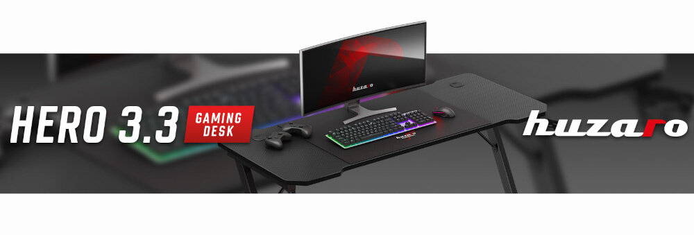 Obraz przedstawia czarne biurko gamingowe z monitorem, klawiaturą i kontrolerem. Na biurku widoczny jest napis \'HERO 3.3 GAMING DESK\' oraz logo \'huzaro\'.