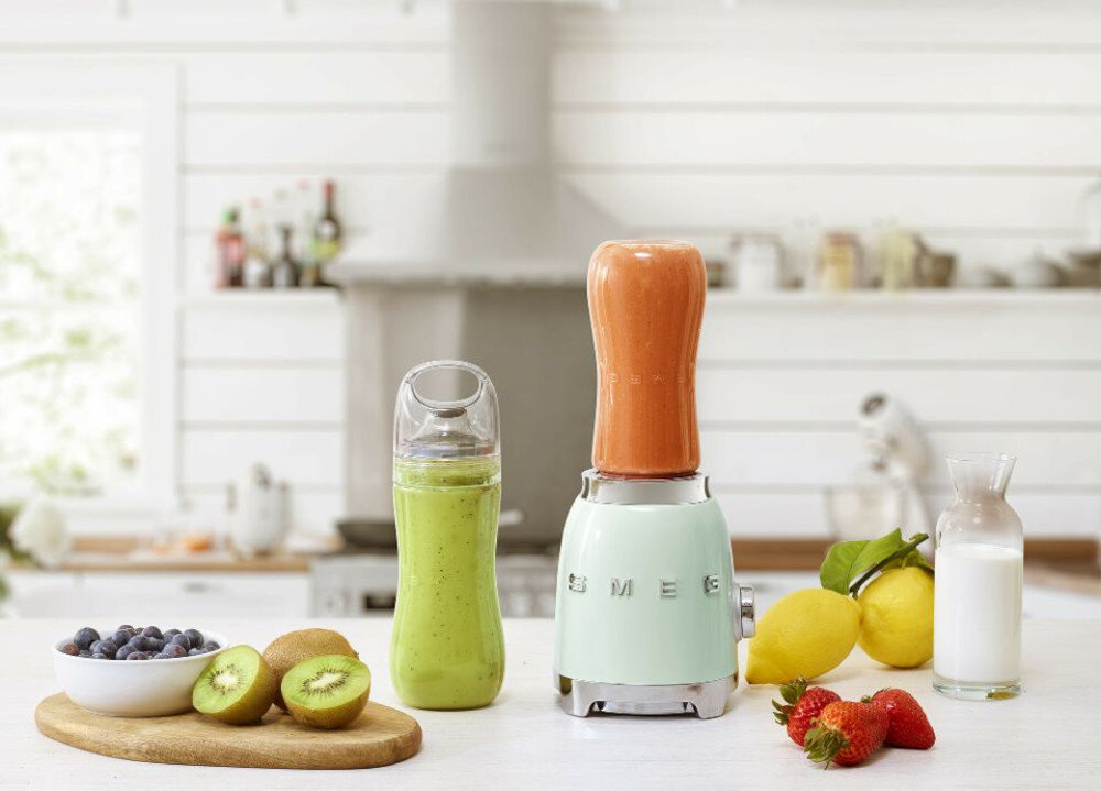 Na blacie kuchennym stoi blender SMEG z różowym koktajlem oraz butelka z zielonym napojem. Obok znajdują się owoce: kiwi, cytryny, truskawki i jagody, a także szklanka mleka.