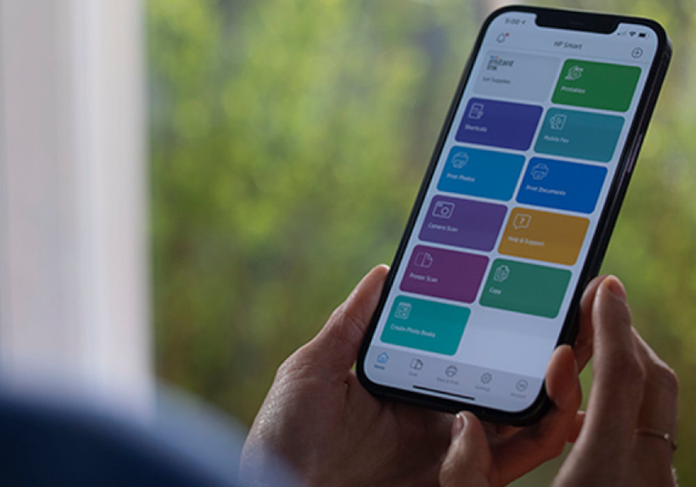 Osoba trzyma smartfon z otwartą aplikacją HP Smart, na której widoczne są kolorowe kafelki z różnymi funkcjami. W tle znajduje się rozmyte zielone tło.