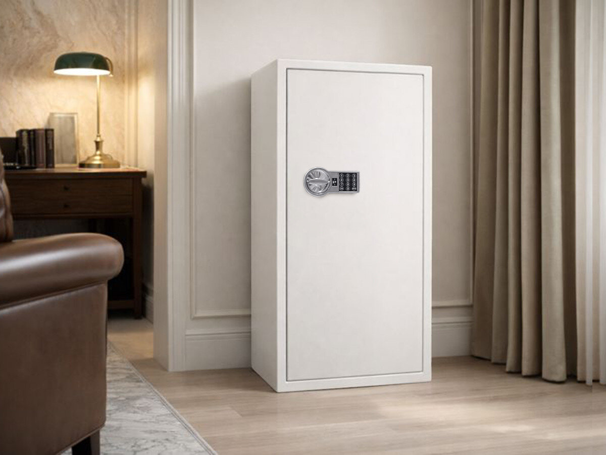 Na obrazie znajduje się biały sejf z elektronicznym zamkiem, umieszczony w eleganckim wnętrzu z zasłonami i biurkiem w tle. Sejf stoi na drewnianej podłodze przy ścianie.