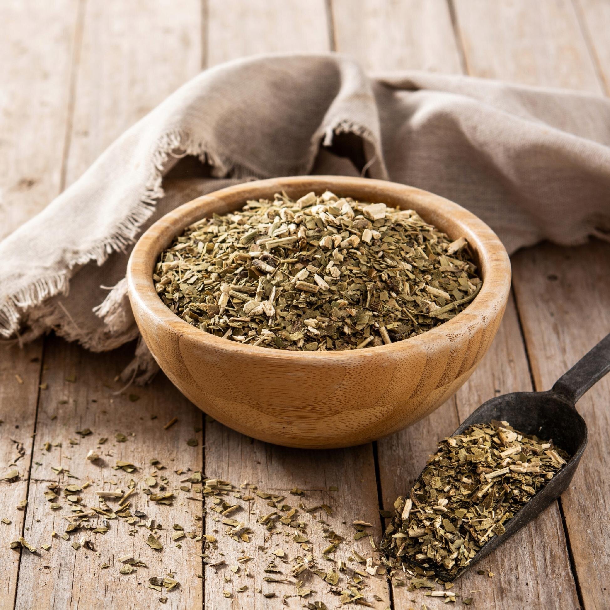 Drewniana miska wypełniona suszem yerba mate stoi na drewnianym stole, obok leży metalowa łopatka z rozsypanym suszem. W tle widoczna jest złożona tkanina.