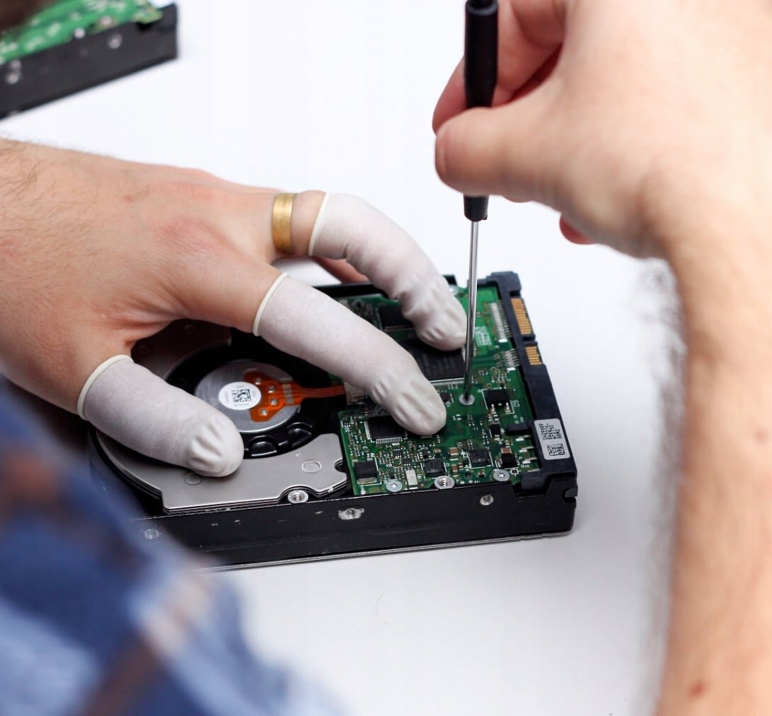 Osoba w lateksowych ochraniaczach na palce używa śrubokręta do pracy nad otwartym dyskiem twardym. W tle widoczna jest część innego urządzenia elektronicznego.