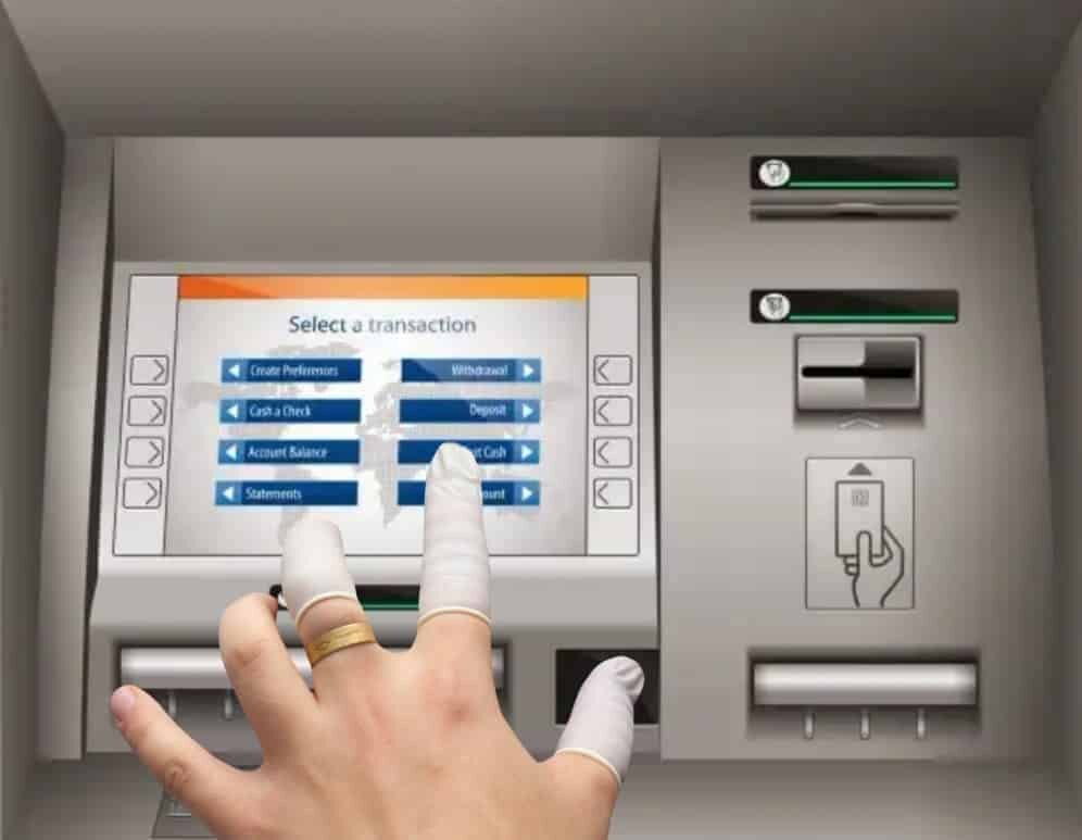 Ręka z założonymi lateksowymi ochraniaczami na palce dotyka ekranu bankomatu. Na ekranie widoczny jest tekst: \'Select a transaction\' z opcjami takimi jak \'Create Preferences\', \'Withdrawal\', \'Cash a Check\', \'Deposit\', \'Account Balance\', \'Transfer Cash\', \'Statements\'.