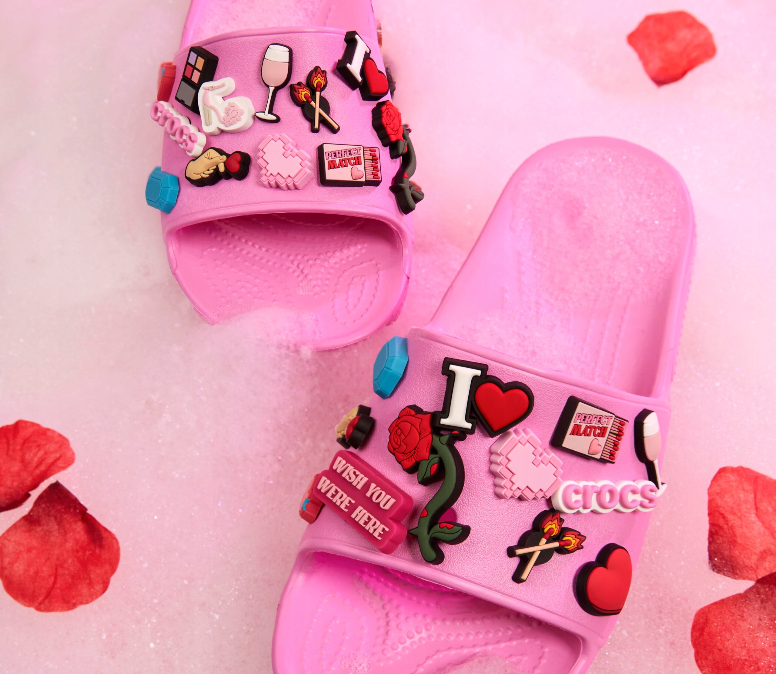 Różowe klapki ozdobione różnorodnymi przypinkami, w tym napisami \'I ❤️\', \'PERFECT MATCH\', \'CROCS\' oraz \'WISH YOU WERE HERE\'. Klapki leżą na różowym tle z płatkami róż.
