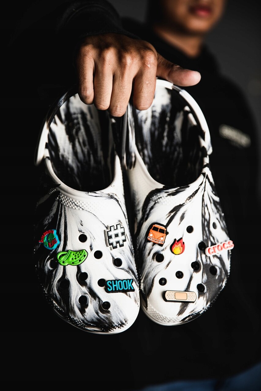 Para białych chodaków z czarnymi wzorami, ozdobionych kolorowymi przypinkami, w tym z napisem \'SHOOK\' i logo \'crocs\'. Buty są trzymane przez osobę w ciemnym ubraniu.