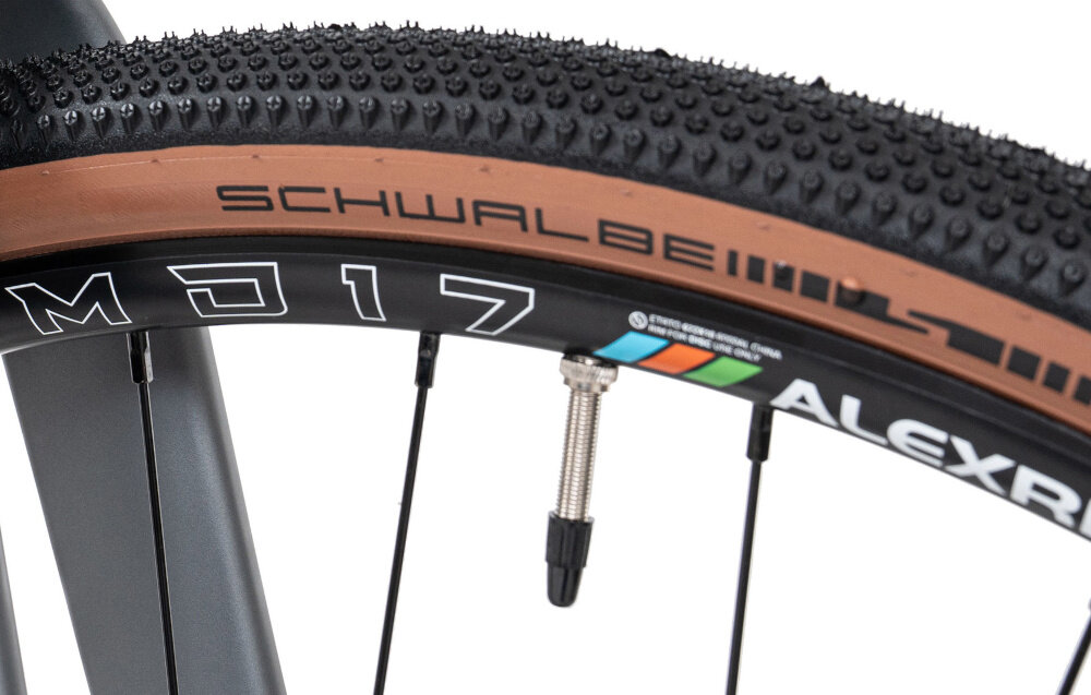 Rower gravel ROMET Aspre 2.0 28540054-A U22 28 cali Oliwkowy zbliżenie na przednie koło białe tło opony marki SCHWALBE w rozmiarze 28 cali głęboki bieżnik skutecznie wspiera jazdę stabilny tor jazdy aluminiowe obręcze sztywność niska masa piasty ROMET D981-12 z przodu i ROMET D982TSE-X12 z tyłu sztywność kół regularne sprawdzanie ciśnienia powietrza wartość na bocznej części opony