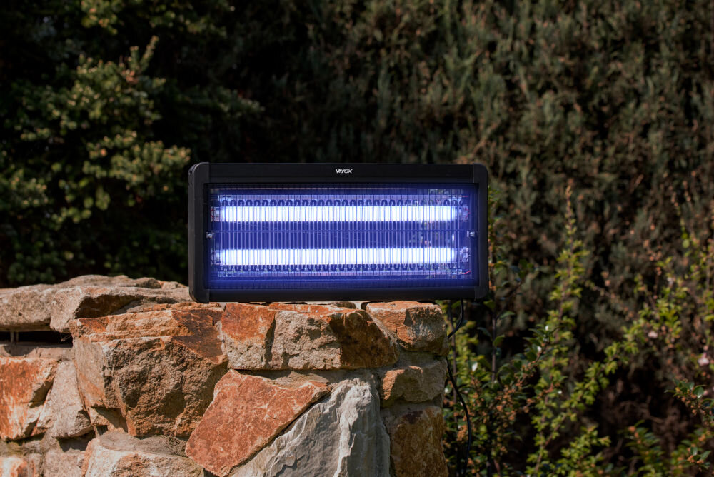 Lampa owadobójcza z napisem \'Vayox\' umieszczona na kamiennej powierzchni, świeci jasnym niebieskim światłem. W tle widoczne są zielone krzewy.