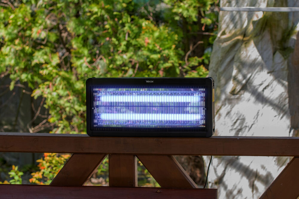 Na drewnianej balustradzie stoi lampa owadobójcza z widocznymi dwoma świetlówkami emitującymi niebieskie światło. Na obudowie lampy widnieje logo \'Vayox\'.