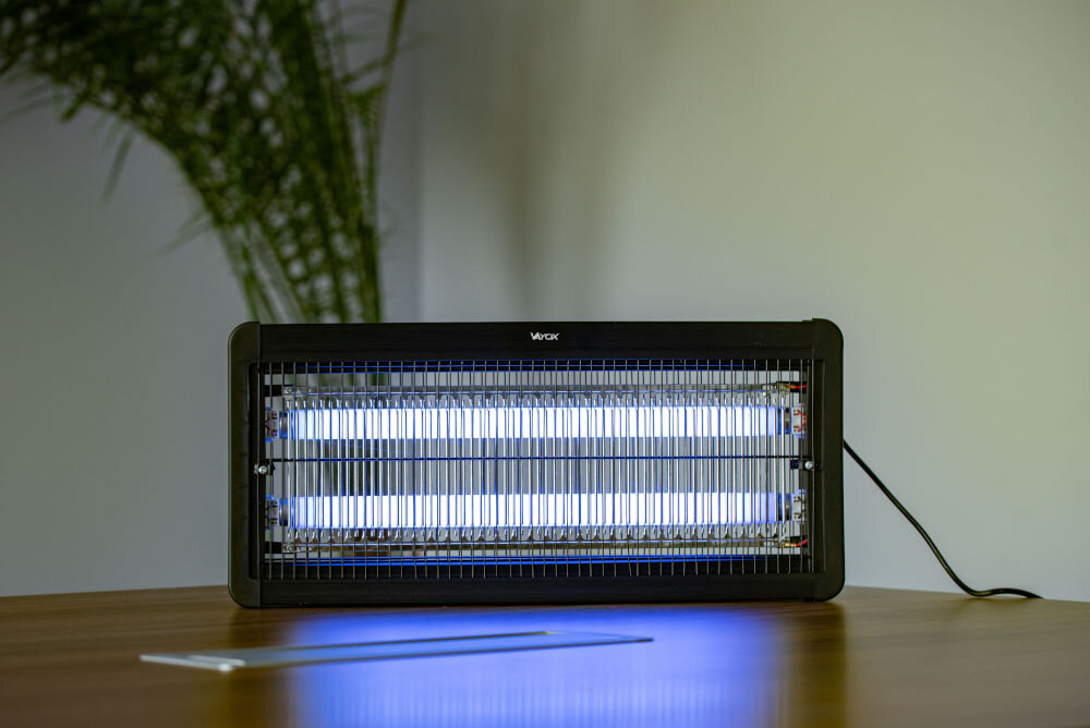 Na drewnianym stole stoi lampa owadobójcza z napisem \'Vayox\', emitująca niebieskie światło. W tle widoczna jest roślina.
