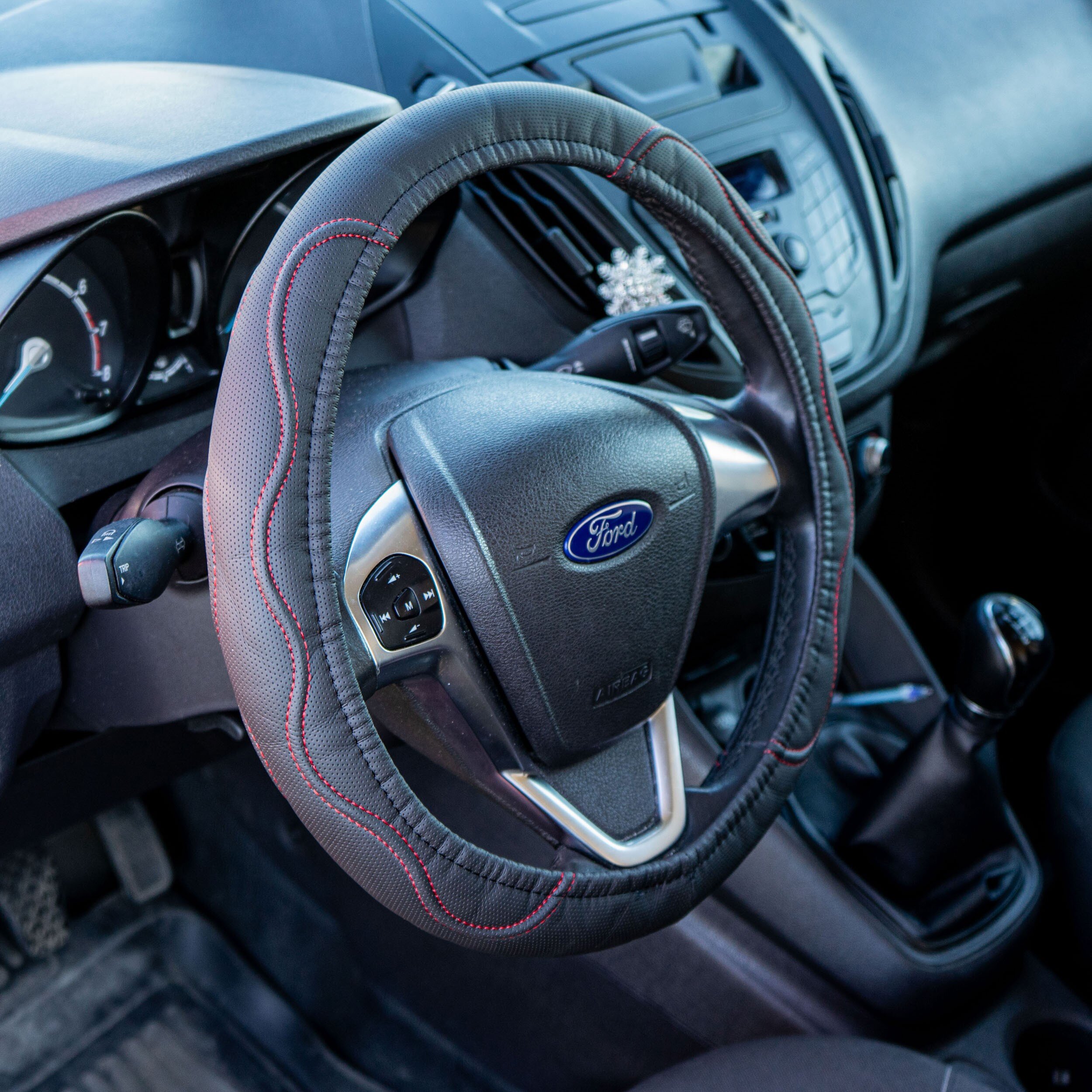 Wnętrze samochodu z widoczną kierownicą, na której znajduje się czarny pokrowiec z czerwonymi przeszyciami. Na środku kierownicy widoczne jest logo Ford.