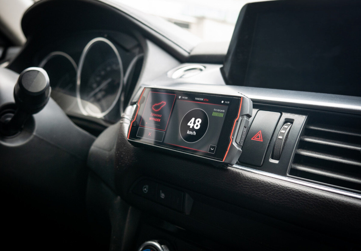 Wnętrze samochodu z zamontowanym na desce rozdzielczej urządzeniem elektronicznym wyświetlającym prędkość 48 km/h. Obok urządzenia widoczny jest przycisk awaryjny i kratka wentylacyjna.