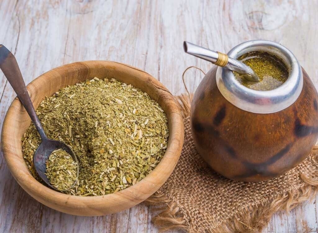 Na drewnianym stole znajduje się miska z suszem yerba mate oraz tykwa z metalową bombillą. Obok leży łyżka zanurzona w suszu.