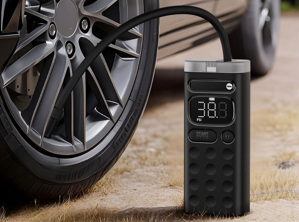 Na obrazie widoczna jest pompka elektryczna z wyświetlaczem pokazującym \'38.8 PSI\', podłączona do opony samochodowej. Pompka ma czarną obudowę z okrągłymi wypustkami i przyciskiem \'START/STOP\'.
