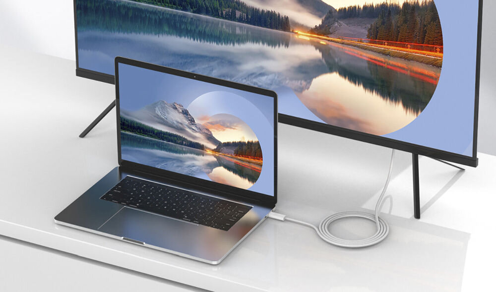 Na białym stole znajduje się laptop podłączony kablem do dużego monitora. Oba ekrany wyświetlają ten sam krajobraz z górami i jeziorem.