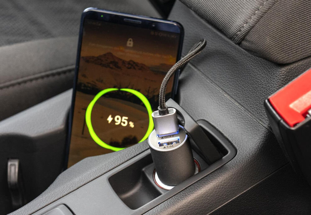 W samochodowym gnieździe zapalniczki znajduje się szara ładowarka z dwoma portami USB, do której podłączony jest kabel. Obok ładowarki stoi telefon z wyświetlonym wskaźnikiem naładowania baterii na poziomie 95%.