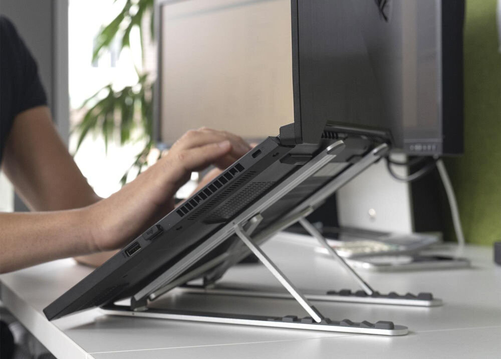 Osoba korzysta z laptopa umieszczonego na metalowej podstawce, która unosi urządzenie pod kątem. W tle widać monitor i roślinę.