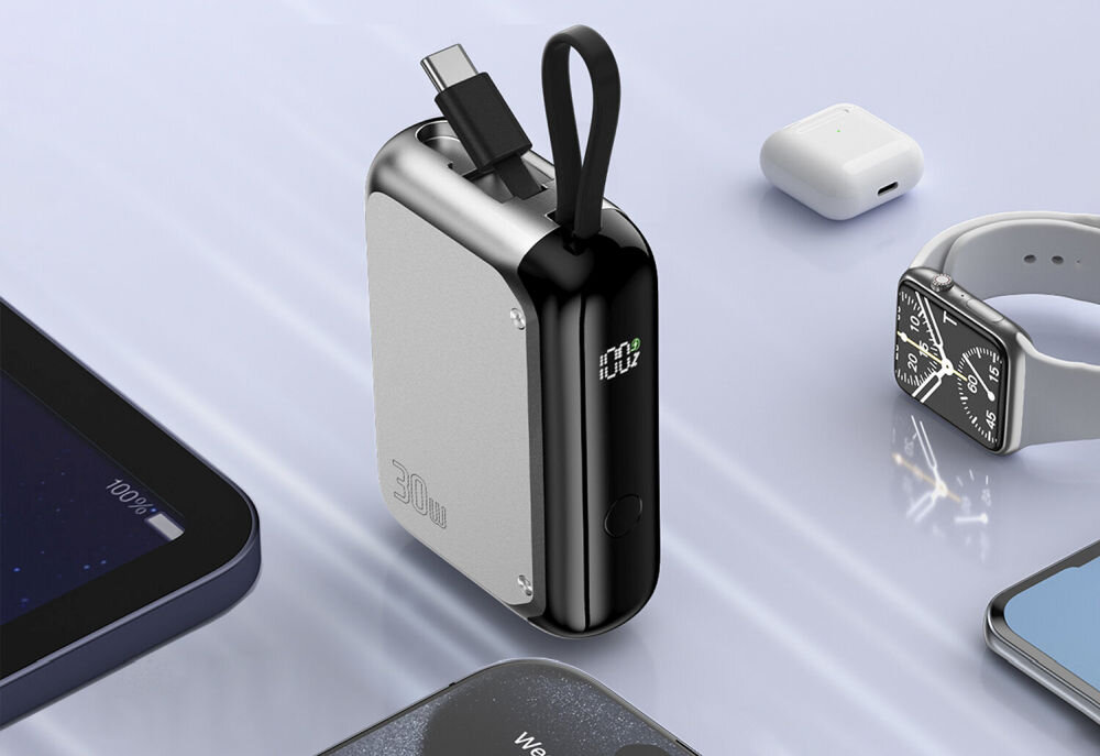 Na obrazie widoczny jest srebrny powerbank z wyświetlaczem LED pokazującym poziom naładowania. Wokół niego znajdują się różne urządzenia elektroniczne, takie jak zegarek, słuchawki i smartfon.