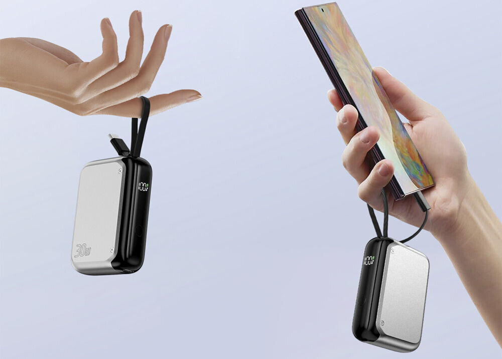 Na obrazie widoczny jest srebrny powerbank z czarnymi bokami, trzymany przez rękę. Powerbank jest podłączony do smartfona, a na jego powierzchni widnieje logo \'4SMARTS\'.