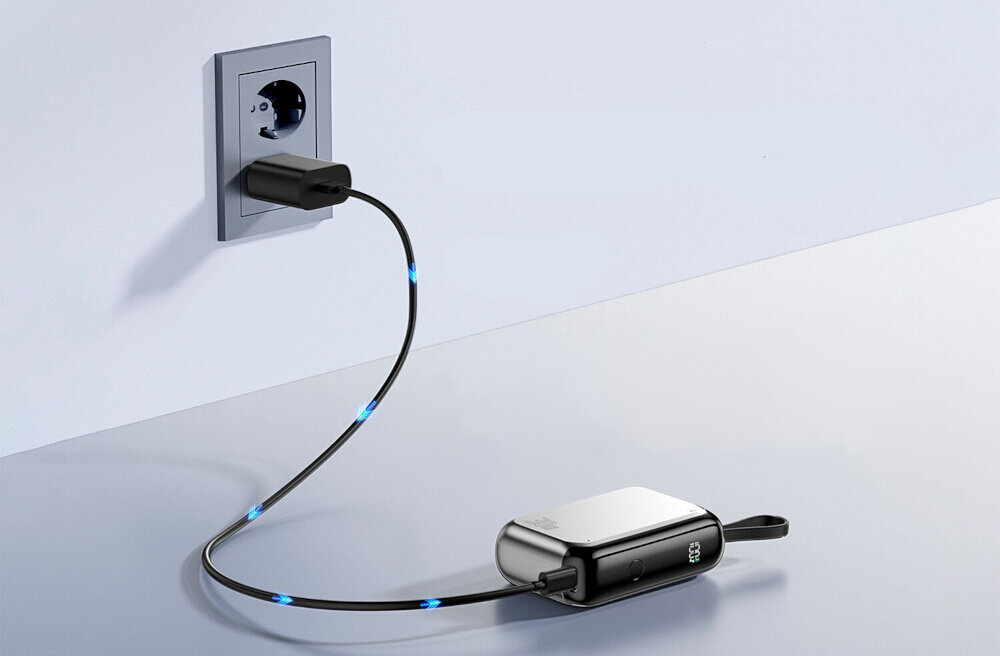 Na obrazie widać powerbank podłączony do gniazdka elektrycznego za pomocą kabla z niebieskimi diodami. Powerbank ma srebrną obudowę i leży na jasnej powierzchni.