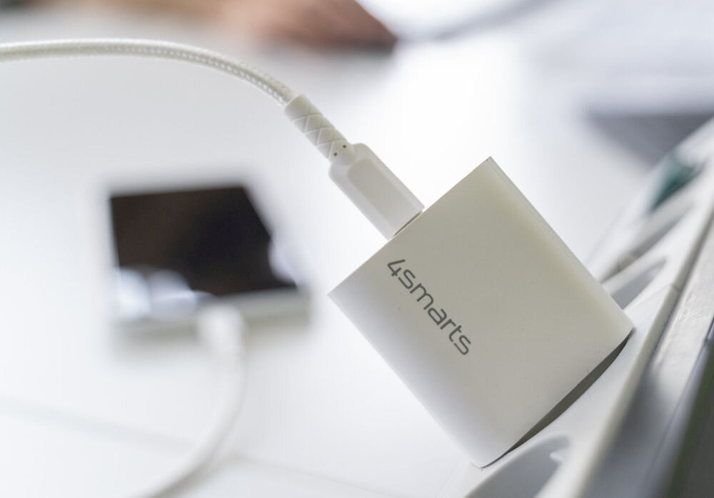 Biała ładowarka z logo \'4smarts\' podłączona do gniazdka, z kablem USB-C. W tle widoczny jest rozmyty telefon.