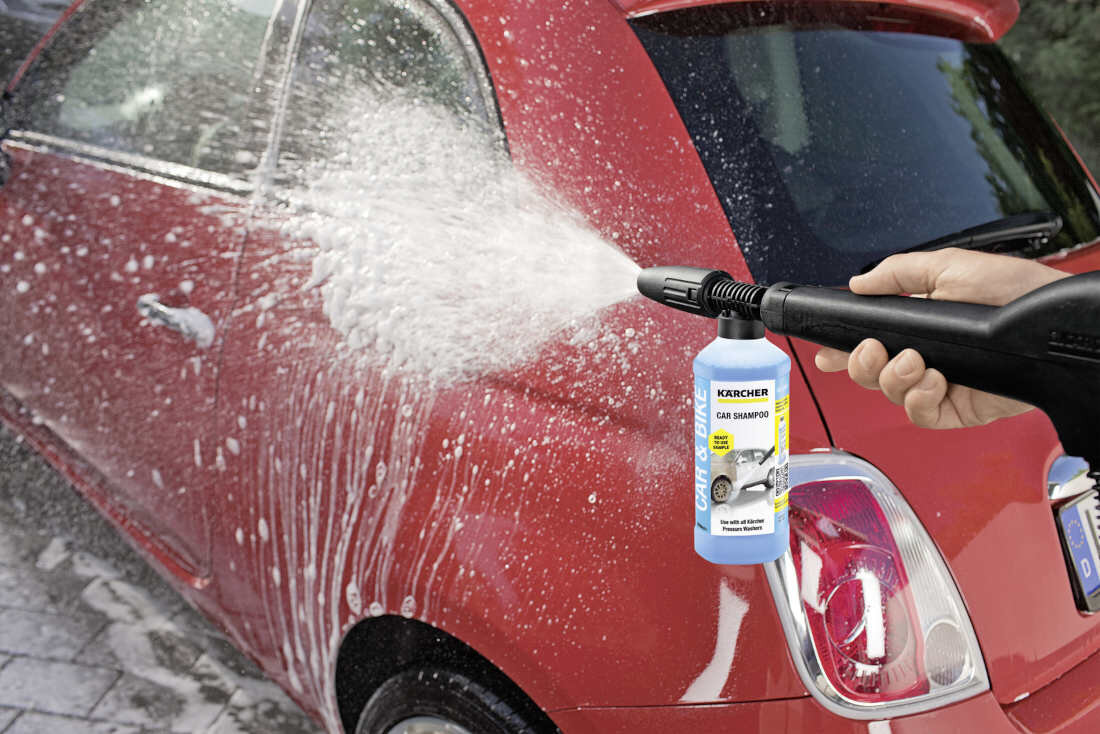 Osoba używa myjki ciśnieniowej do czyszczenia czerwonego samochodu. Na butelce widoczny jest napis \'KARCHER Car Shampoo\'.