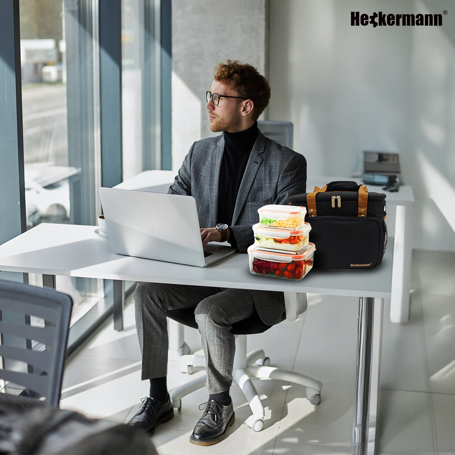 Mężczyzna siedzi przy biurku z laptopem, obok niego stoi czarna torba termiczna Heckermann oraz pojemniki z jedzeniem. W tle widoczne jest logo Heckermann.
