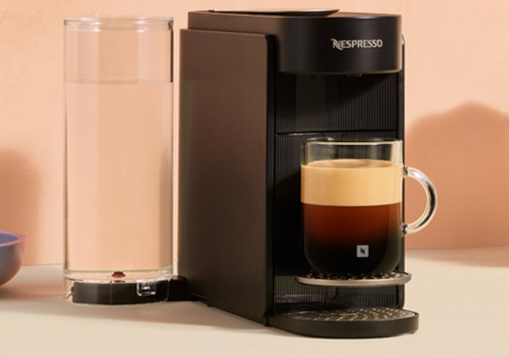 Na obrazie widoczny jest czarny ekspres do kawy z logo \'Nespresso\', obok którego stoi przezroczysty pojemnik na wodę. Na ekspresie znajduje się szklanka z kawą z pianką.