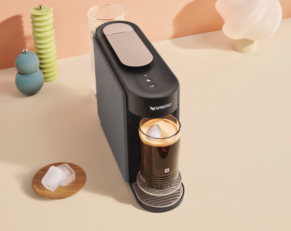 Na blacie stoi ekspres do kawy z logo Nespresso, z przygotowanym napojem w szklance. Obok znajduje się talerzyk z kostkami lodu.