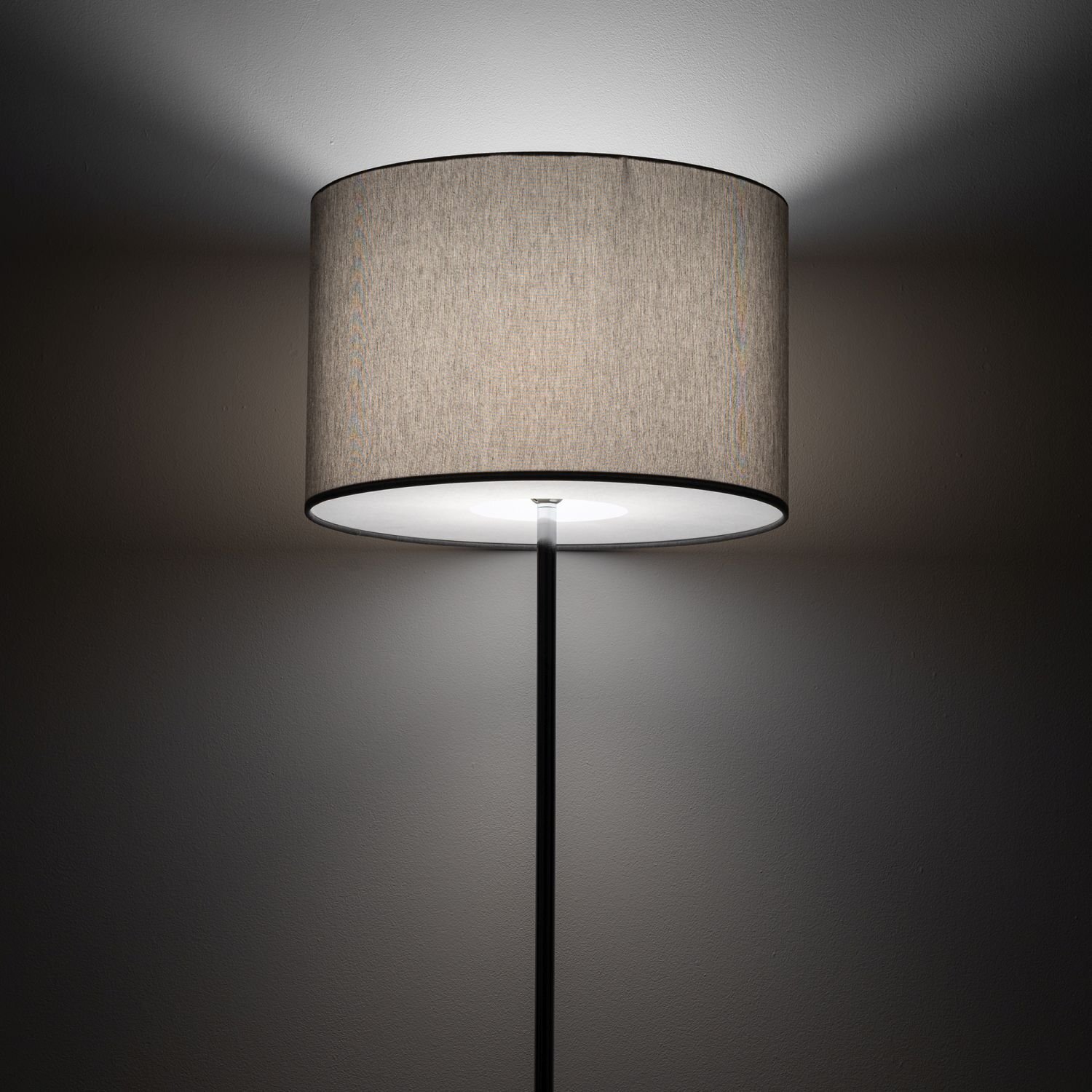 Lampa podłogowa z okrągłym, beżowym abażurem, emitująca światło na ścianę. Czarny, prosty trzon lampy prowadzi do abażura.