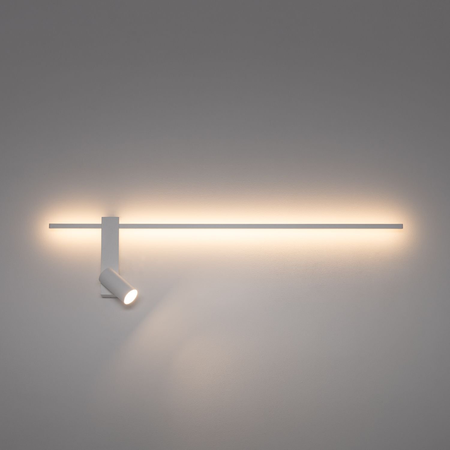 Na obrazie widoczna jest nowoczesna lampa ścienna z reflektorem, emitująca ciepłe, rozproszone światło. Lampa ma prosty, minimalistyczny design z długim, poziomym elementem świetlnym i punktowym reflektorem skierowanym w dół.