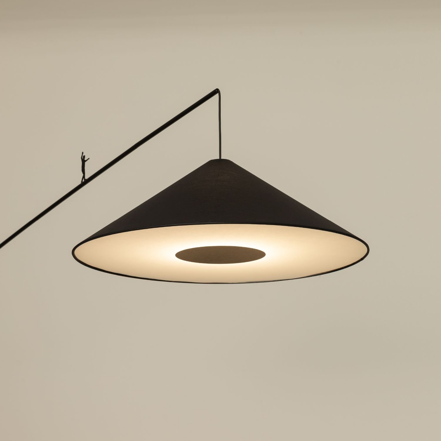 Na obrazie widoczna jest lampa podłogowa z dużym, stożkowym kloszem w kolorze ciemnym, emitująca ciepłe światło. Lampa posiada metalowy wysięgnik z małą figurką na górze.