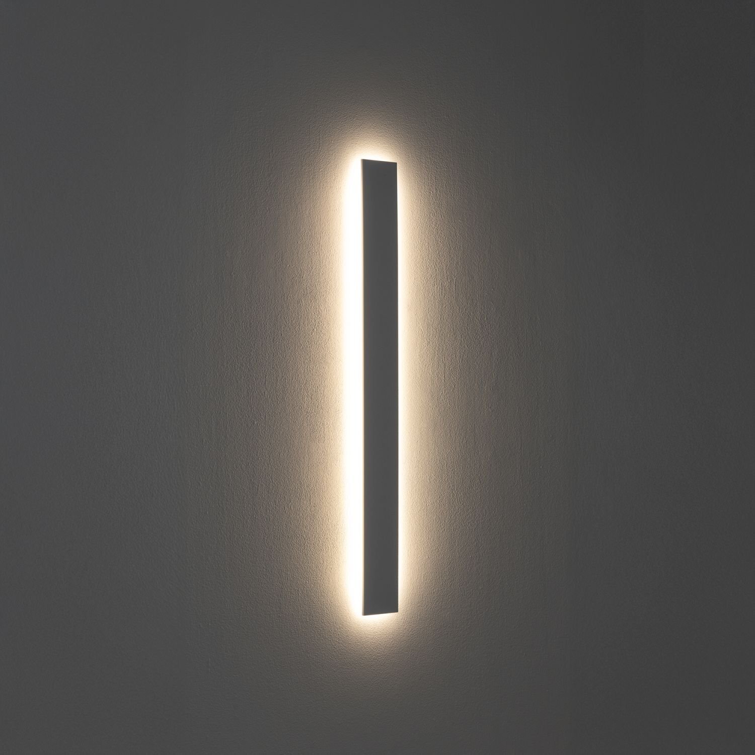 Na obrazie widoczna jest nowoczesna lampa ścienna LED o liniowym kształcie, emitująca delikatne światło na ścianę. Lampa ma prosty, minimalistyczny design.