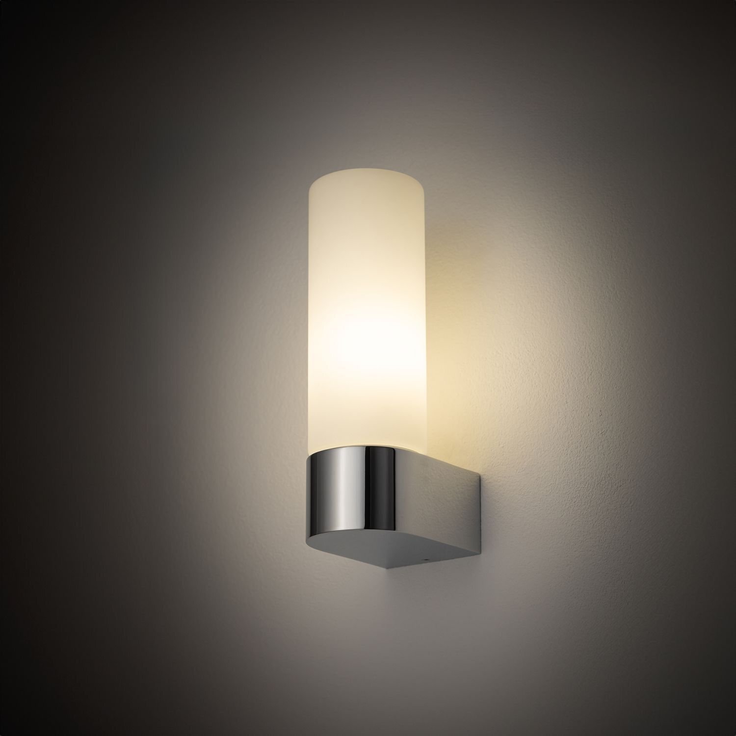 Na obrazie widoczny jest elegancki kinkiet ścienny z cylindrycznym, mlecznym kloszem i metalową podstawą w kolorze srebrnym. Lampa emituje ciepłe, rozproszone światło na ścianę.