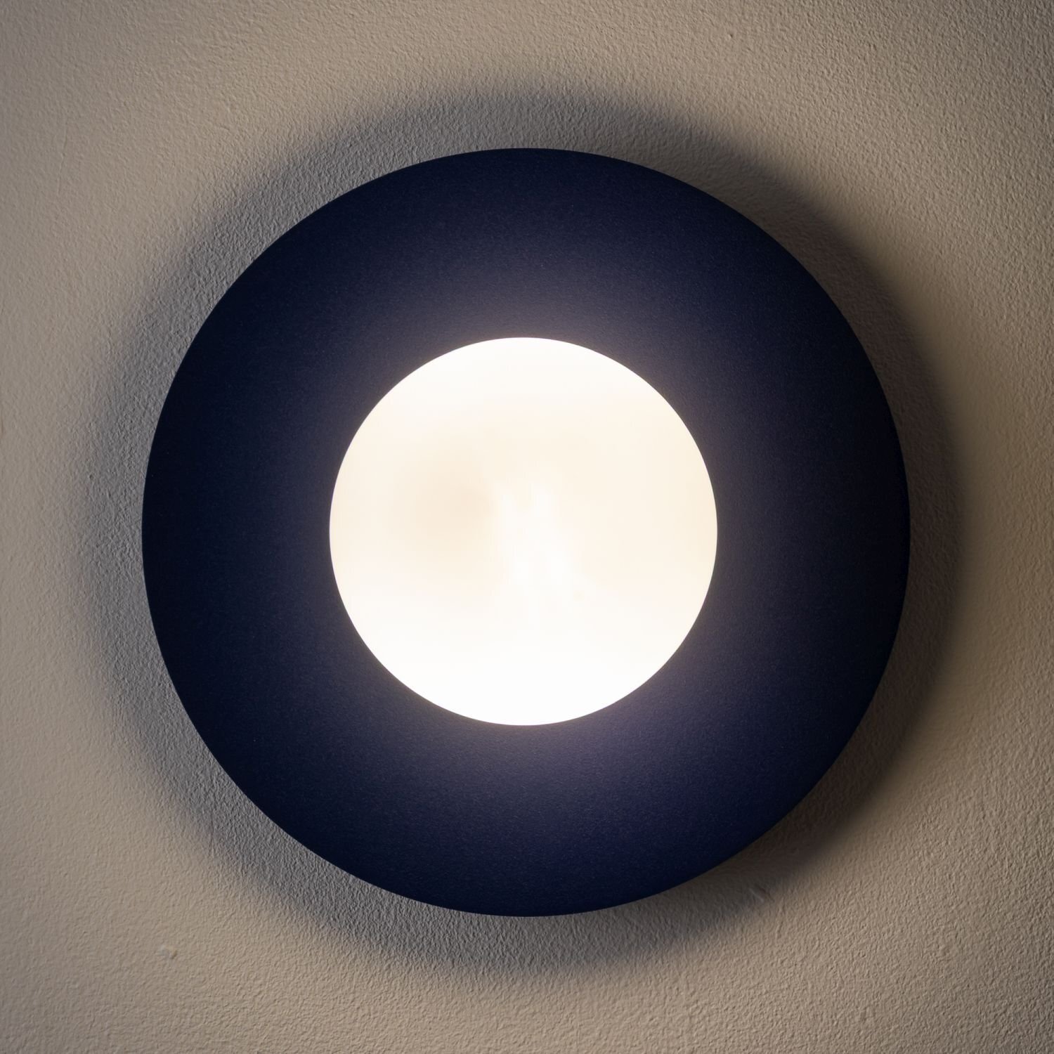 Na obrazie widoczna jest okrągła lampa ścienna z jasnym, centralnym źródłem światła otoczonym ciemnym pierścieniem. Lampa jest zamontowana na ścianie, a światło tworzy delikatny efekt świetlny na powierzchni.