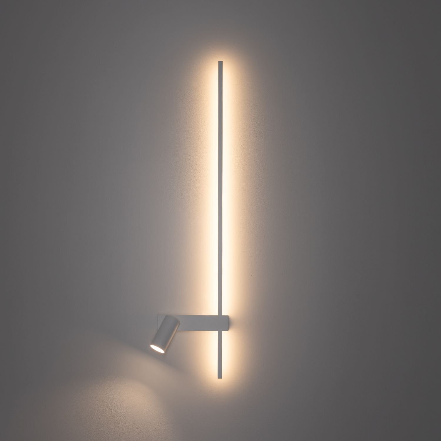 Na obrazie widoczna jest nowoczesna lampa ścienna LED z długim, pionowym paskiem świetlnym oraz reflektorem skierowanym w dół. Lampa emituje ciepłe, rozproszone światło na ścianę.