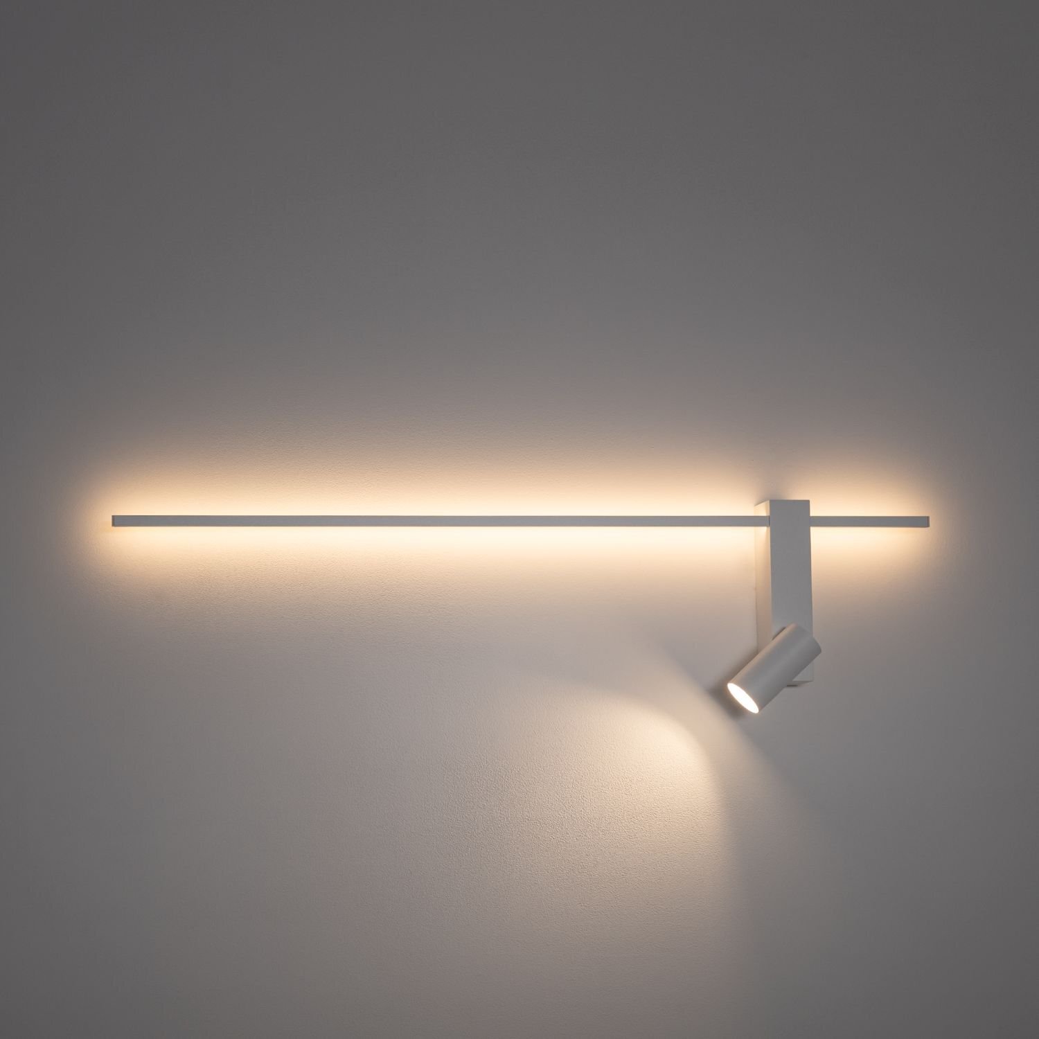 Na obrazie widoczna jest nowoczesna lampa ścienna LED z długim, poziomym panelem świetlnym i reflektorem skierowanym w dół. Lampa emituje ciepłe, rozproszone światło na ścianę.