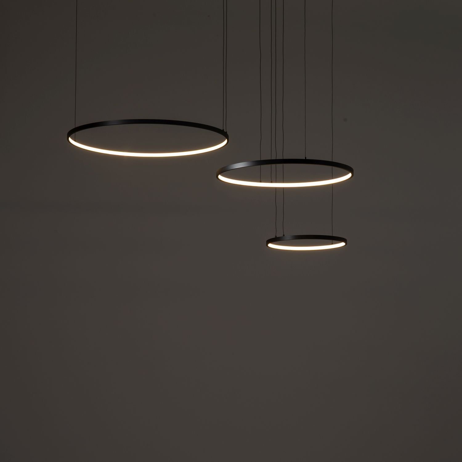 Trzy okrągłe, wiszące lampy LED o nowoczesnym designie, zawieszone na cienkich przewodach. Lampy emitują delikatne, białe światło na ciemnym tle.