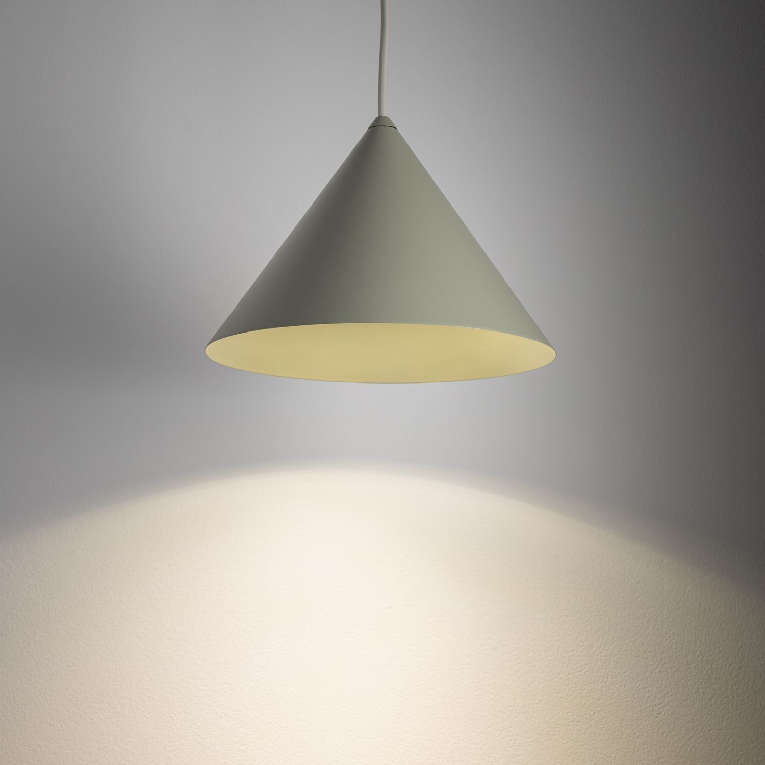 Na obrazie widoczna jest wisząca lampa o stożkowym kształcie, emitująca światło na ścianę. Lampa ma jednolity, jasny kolor.