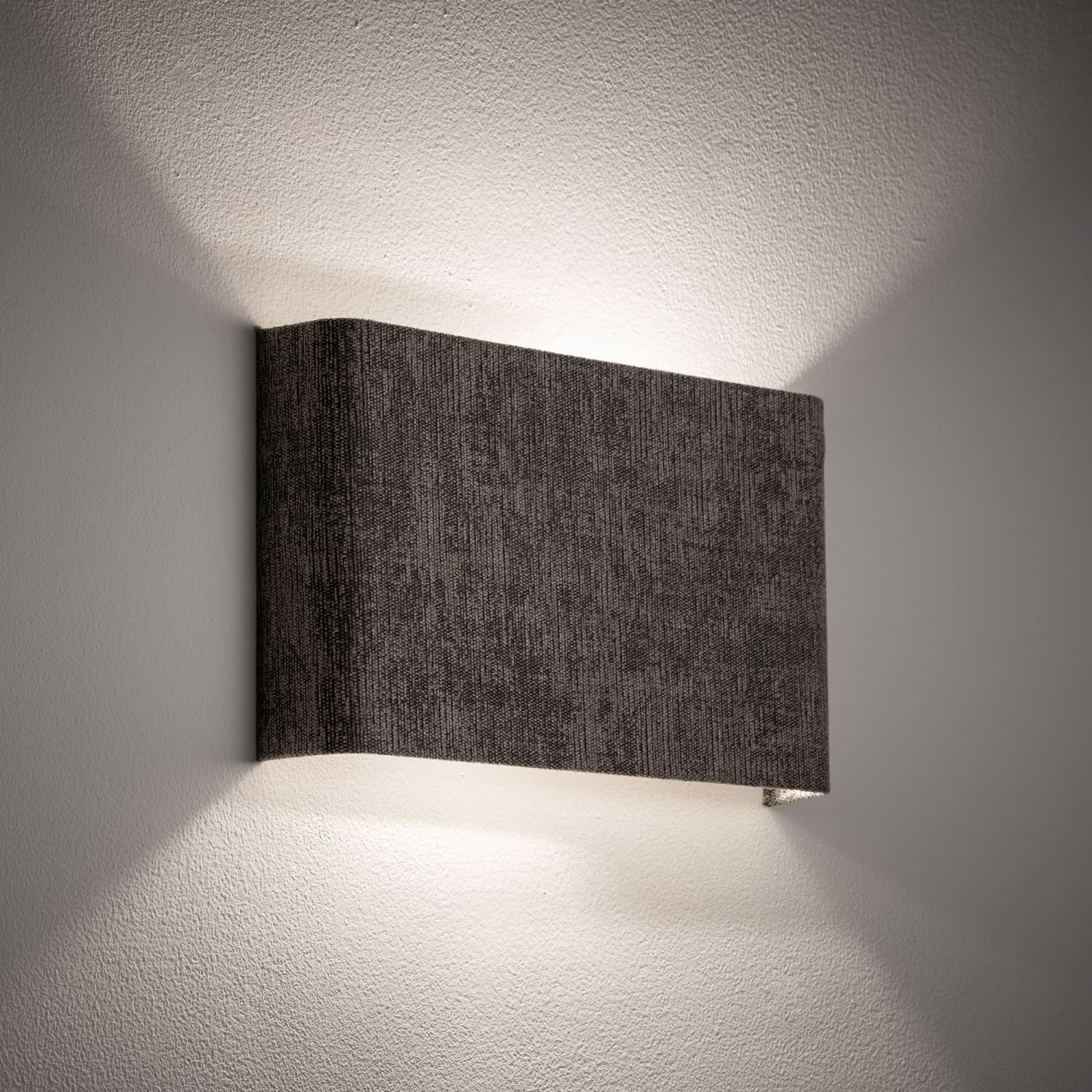Na obrazie widoczna jest nowoczesna lampa ścienna o prostokątnym kształcie, emitująca światło w górę i w dół. Lampa ma ciemną, teksturowaną powierzchnię i jest zamontowana na jasnej ścianie.